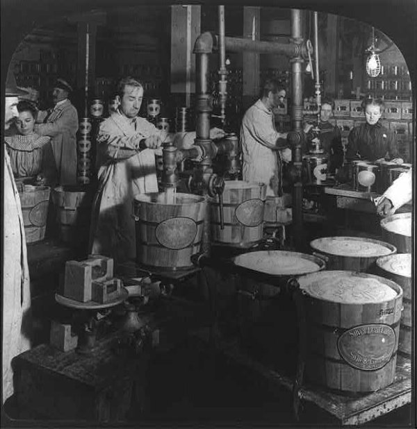 Black and white photo of workers in a meatpacking plant filling large buckets with lard. 