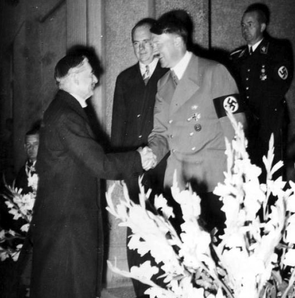 A black-and-white photograph shows Chamberlain and Hitler shaking hands indoors in a formal setting. 