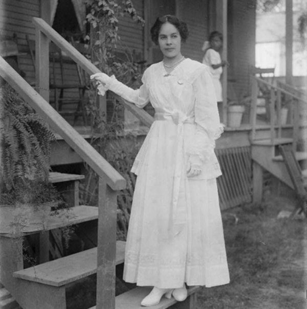 Black and white photograph of a young woman in a long white dress standing on a porch, holding a wooden railing with one hand. 
