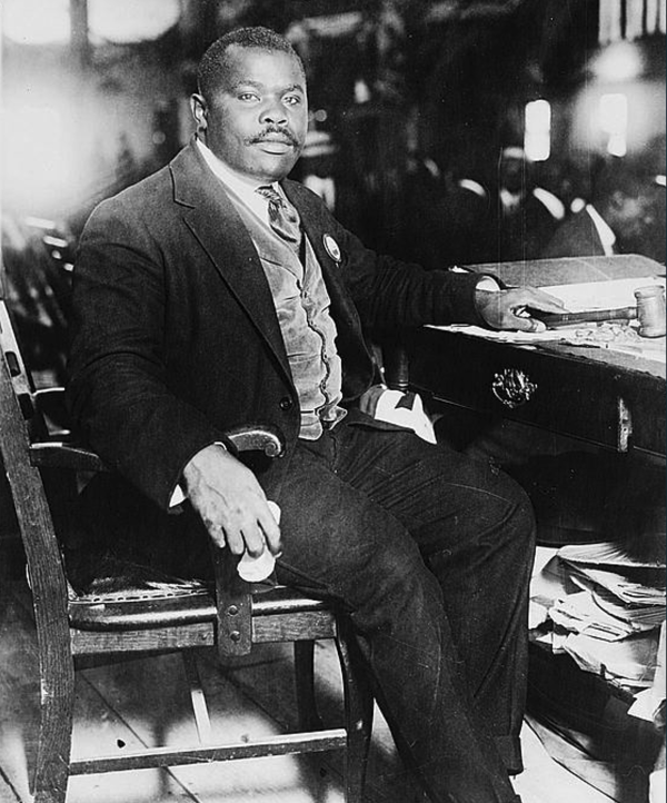 Black and white photograph of Marcus Garvey seated at a desk in a formal setting, wearing a suit and tie. 