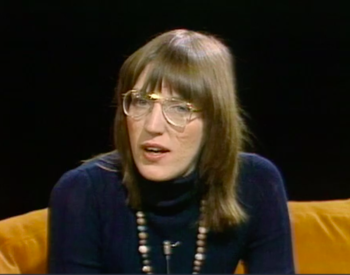 Photo of a woman with brown hair and glasses wearing a navy turtleneck, seated on a yellow couch against a dark background.